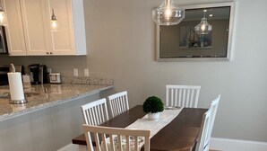 Een eetruimte met een houten tafel en witte stoelen, een vloerkleed met blauw patroon, en een keuken op de achtergrond.