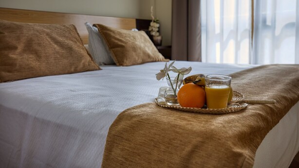 Una cama bien hecha con una bandeja de jugo de naranja y un vaso de jugo, un almohón y una mesita de noche con un jarrón de flores.