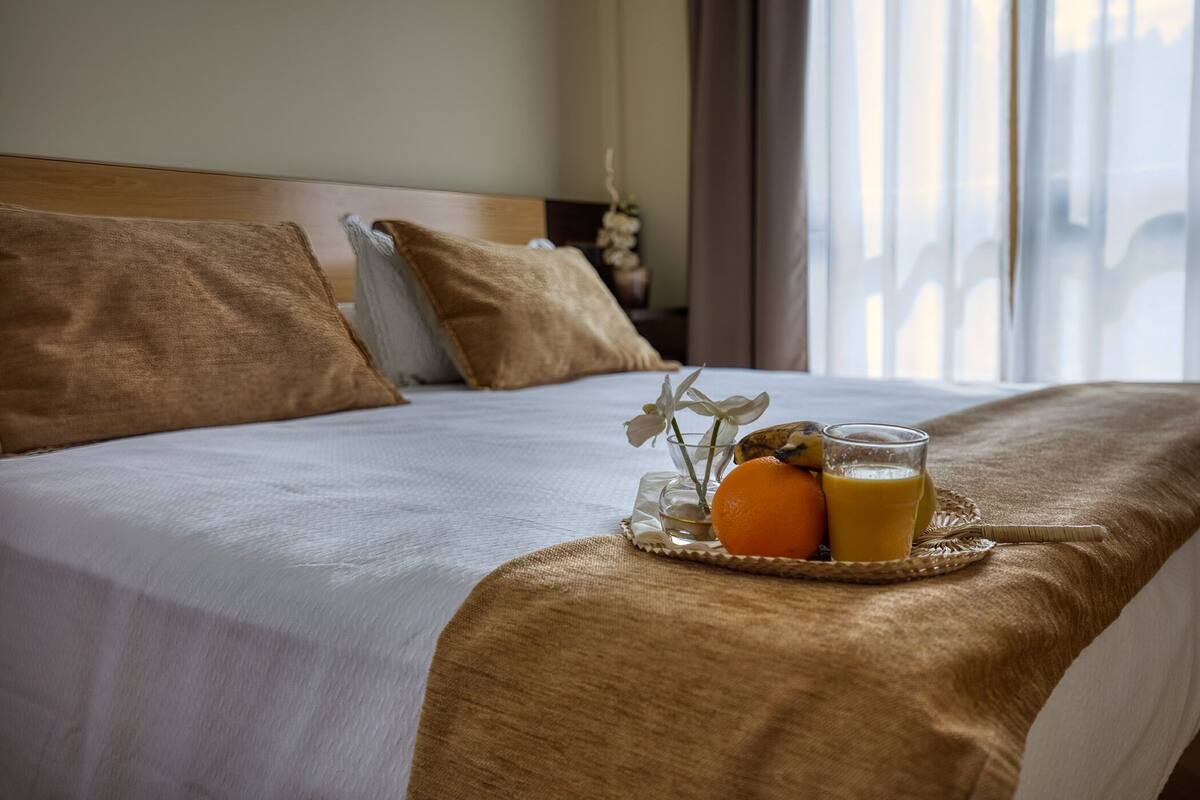 A neatly made bed with a tray of orange juice and a glass of orange juice, a pillow, and a bedside table with a vase of flowers.