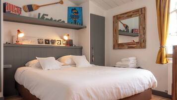 Een net opgemaakt bed met witte lakens, een houten hoofdbord en een plank erboven met decoratieve objecten en een ingelijste foto.