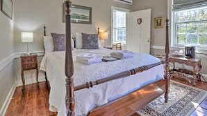 A four-poster bed with a white bedspread, a wooden nightstand with a lamp, a small table with a lamp, and a window with a view of trees. - Historic Warrenton Home w/ Patio & Hot Tub! (Warrenton)