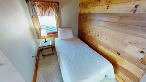 Una habitación pequeña con cama individual, pared de madera, mesita de noche con lámpara y ventana con cortinas.