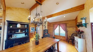 Una mesa de comedor de madera con sillas, una araña y una zona de cocina con paredes de madera y un ventanal con cortinas.