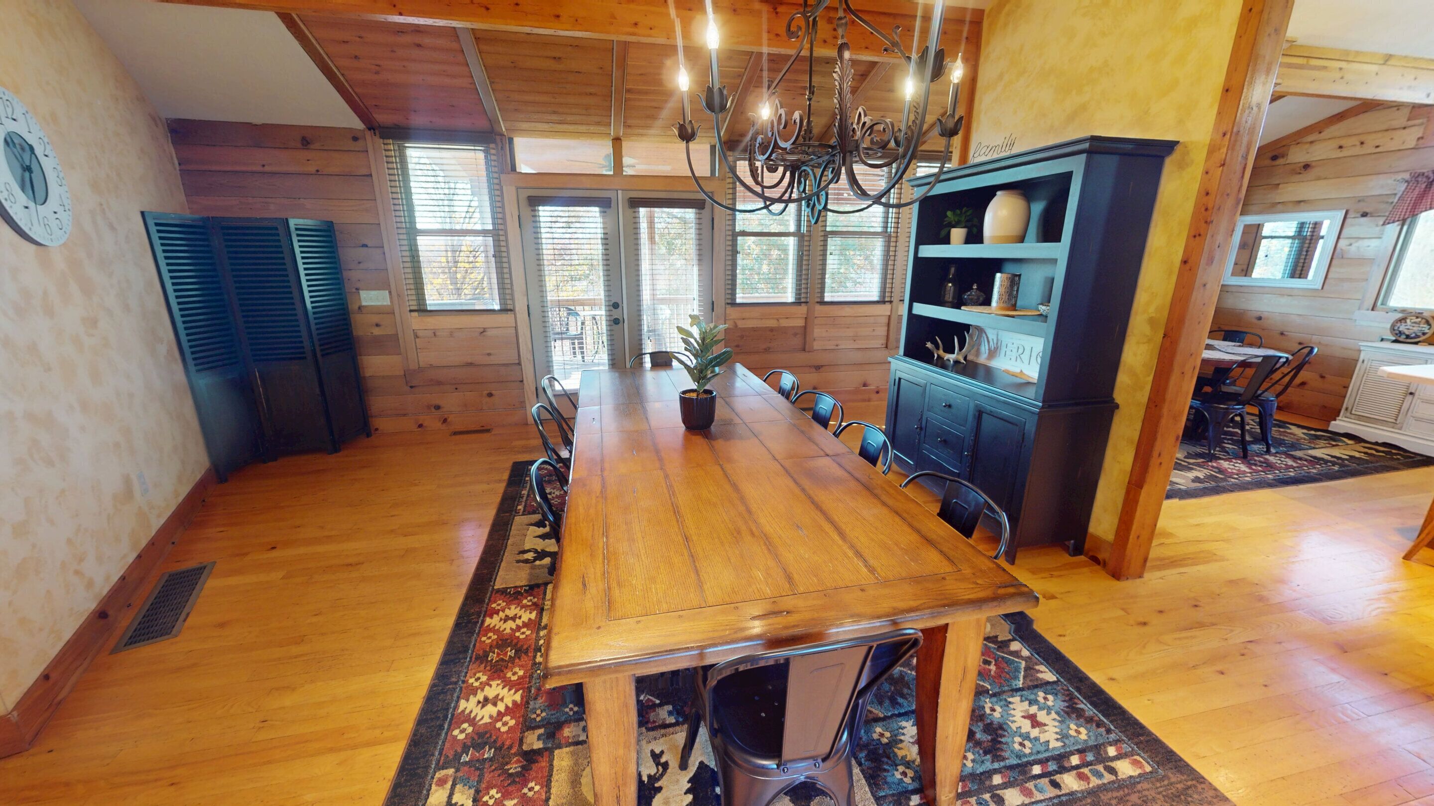 A spacious dining area with a long wooden table, chairs, a chandelier, and a blue cabinet with shelves.