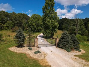 A gravel driveway leading to a gated entrance with a deer symbol, surrounded by trees and a clear sky.