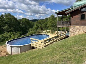 En swimmingpool med træterrasse og stenvæg, omgivet af græs og træer.