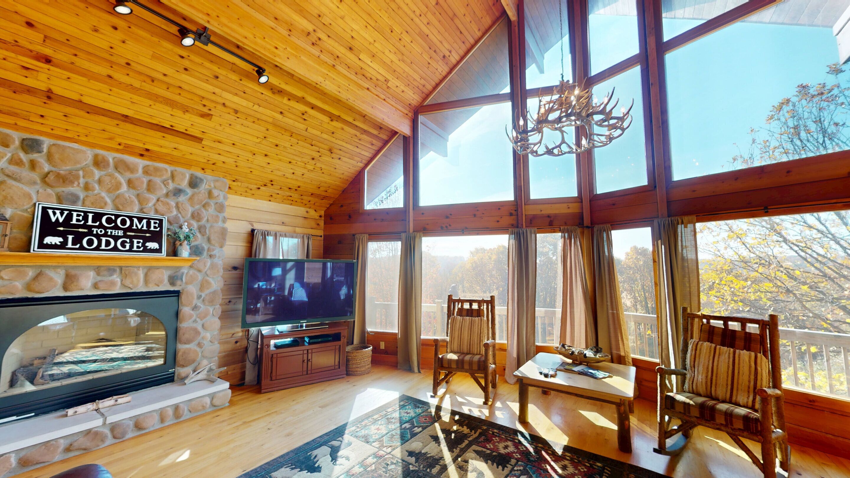 L’intérieur d’un chalet présente une cheminée en pierre, un téléviseur, des meubles en bois et de grandes fenêtres donnant sur la forêt.