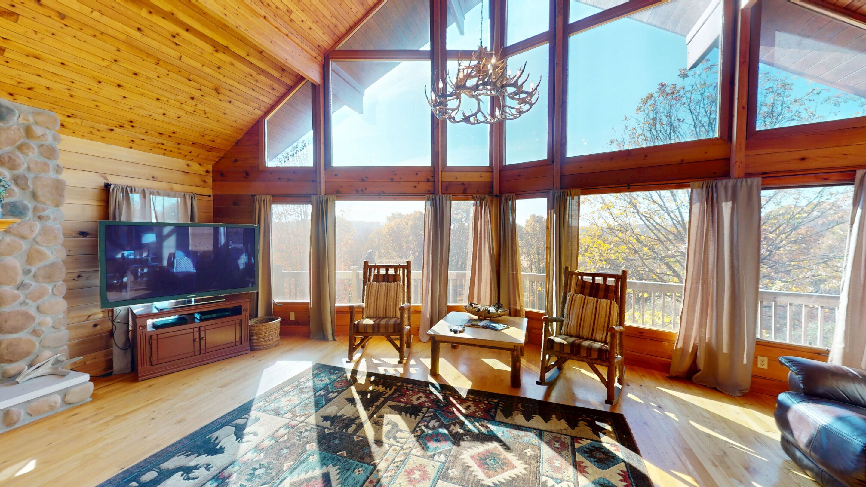 Interior acogedor de una cabaña con chimenea de piedra, televisor, mobiliario de madera y ventanas amplias con vistas al exterior.