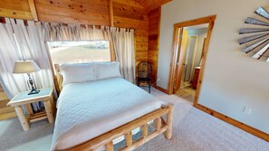 A wooden cabin bedroom with a bed, a nightstand, a chair, and a view of the outdoors.