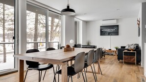 Ein modernes Wohnzimmer mit großem Fernseher, einem Essbereich mit Holztisch und Stühlen sowie Blick durch große Fenster.
