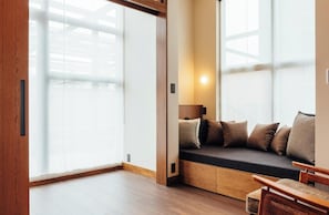 Une chambre d’hôtel moderne dotée d’une porte-fenêtre coulissante, d’une banquette intégrée avec des coussins, d’un plancher en bois et d’une grande fenêtre laissant entrer la lumière naturelle.