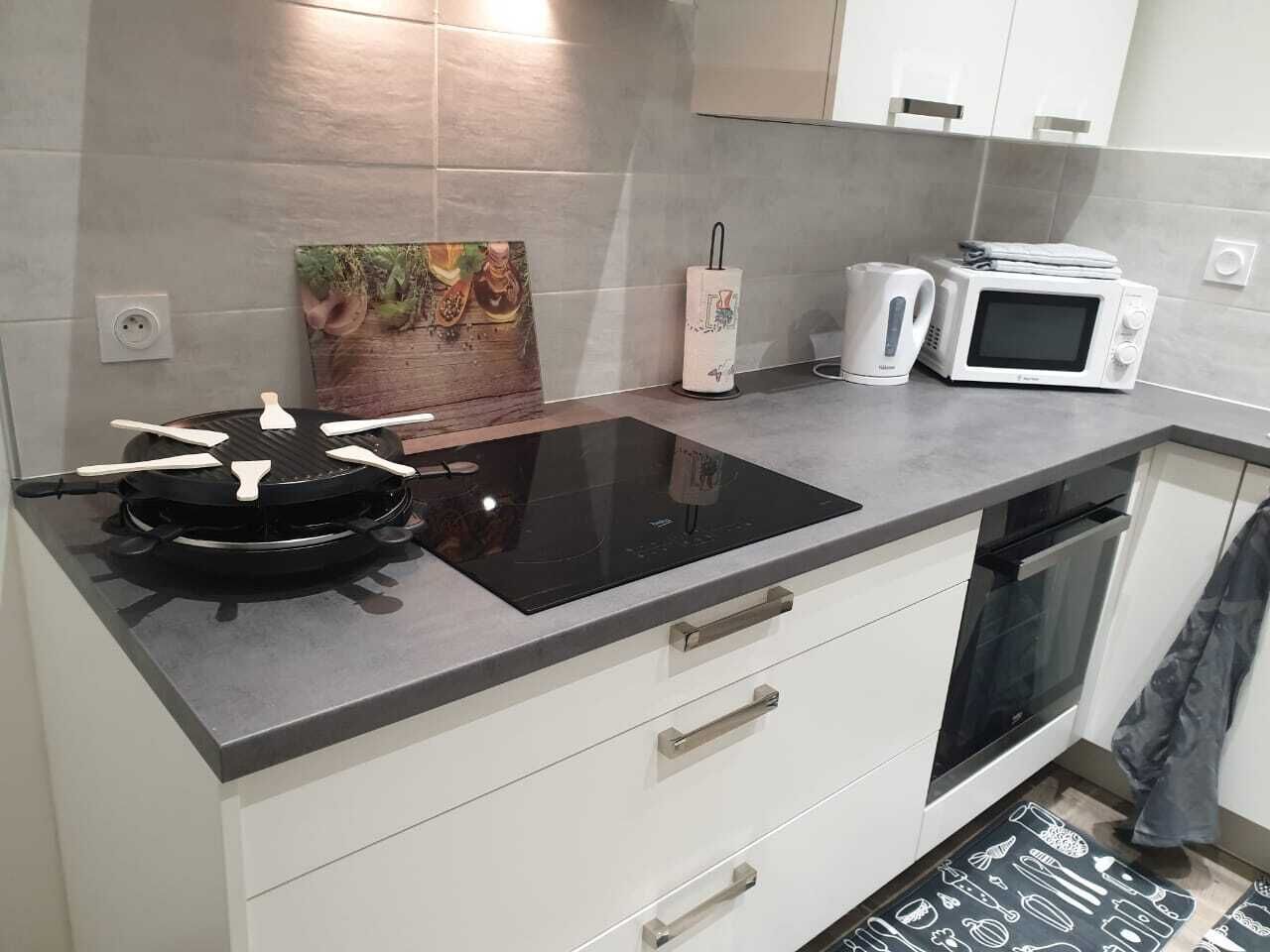 Een keuken met een zwart inductiekookplaatje, een magnetron en een waterkoker. Er hangt een schilderij aan de muur en er ligt een klein vloerkleed op de grond.