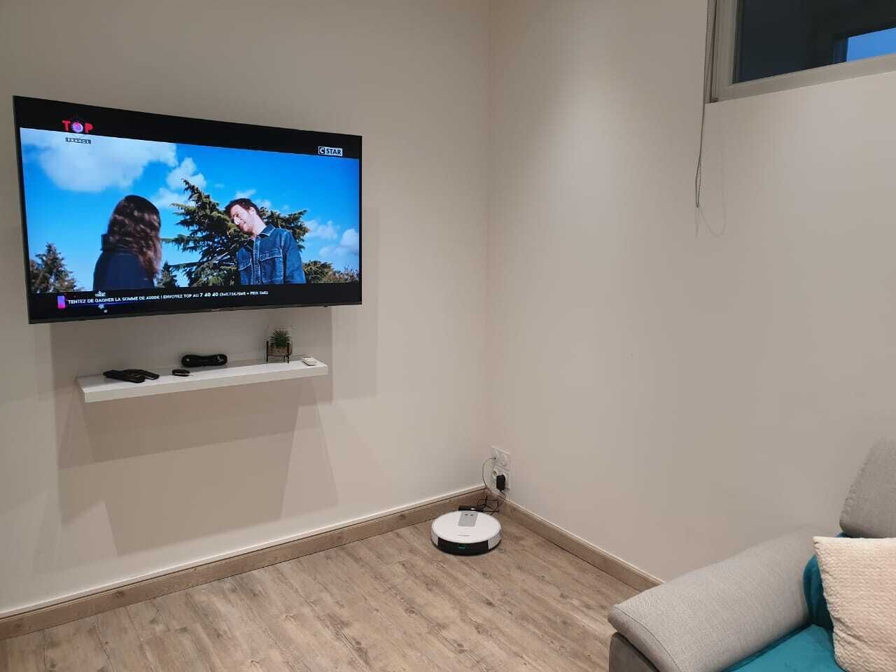 Una stanza con una TV a parete che mostra un programma, uno scaffale bianco con degli oggetti, un aspirapolvere robot e un divano.