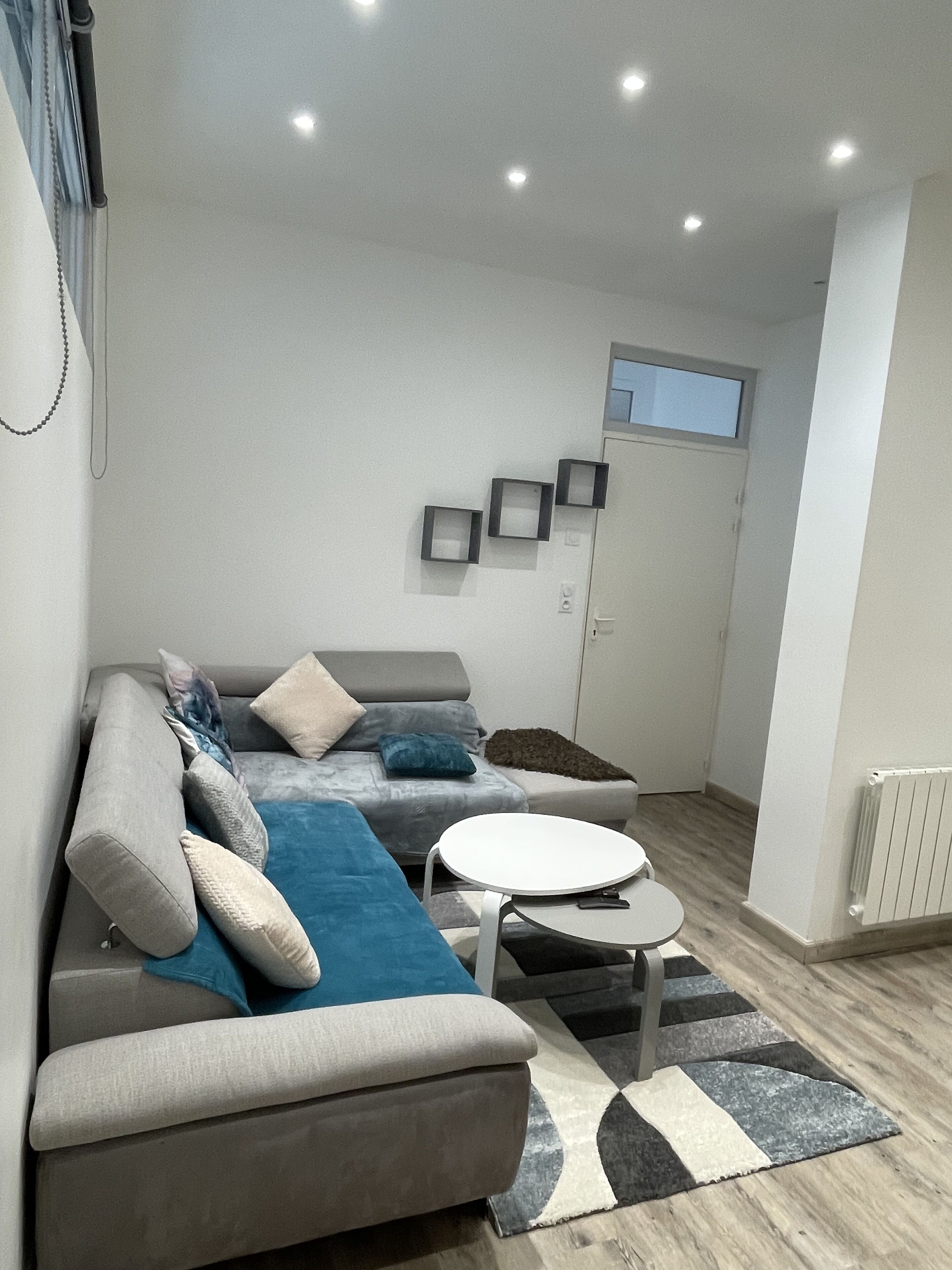 Un salon moderne avec un canapé gris, des coussins bleus et beiges, une table basse blanche et un tapis à motifs. Il y a trois étagères fixées au mur et une fenêtre au-dessus de la porte.