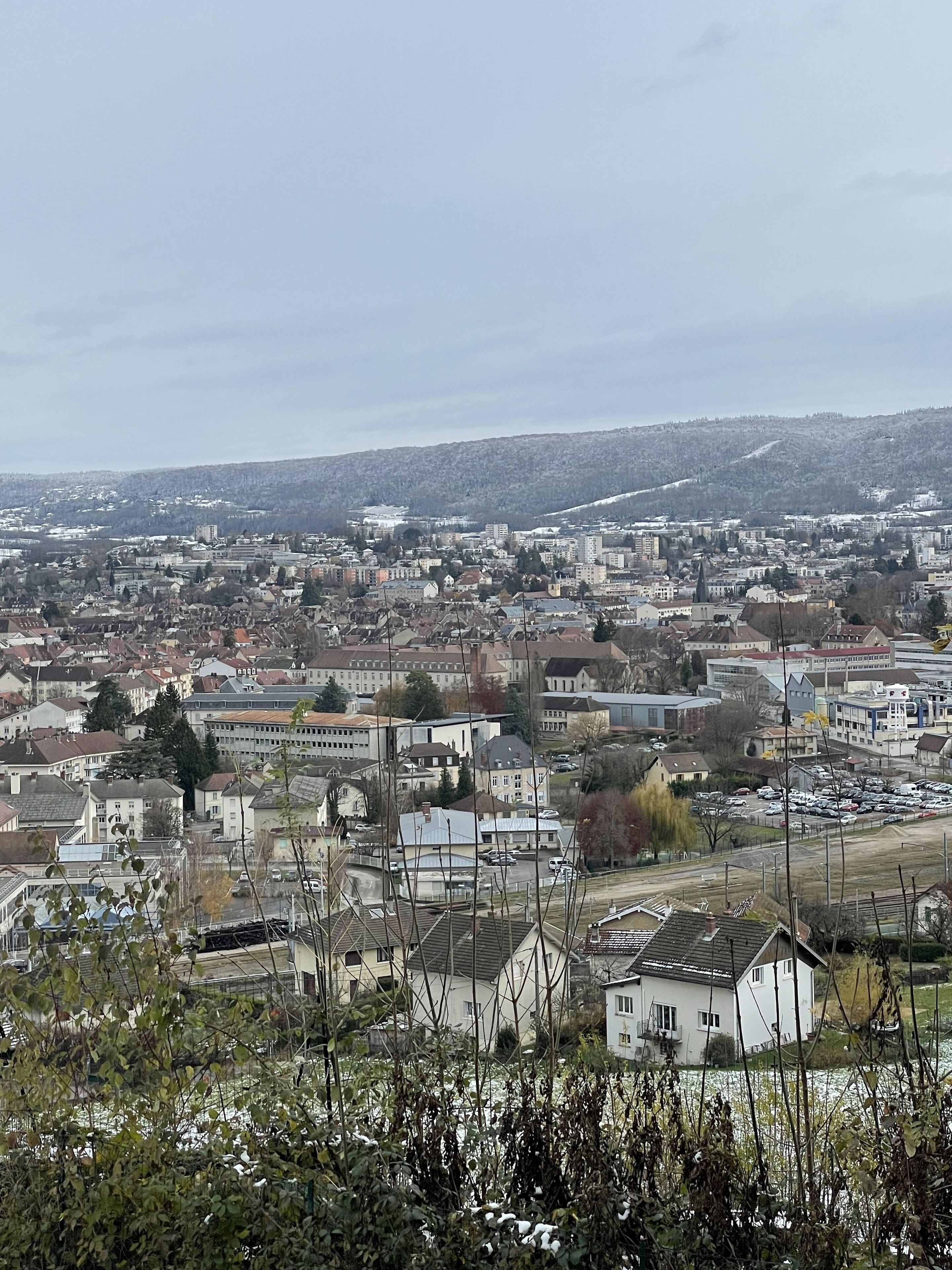 Ein weiter Panoramablick auf eine Stadtlandschaft mit einer Mischung aus Wohn- und Geschäftsgebäuden, einem Fluss im Vordergrund und einer hügeligen Kulisse.