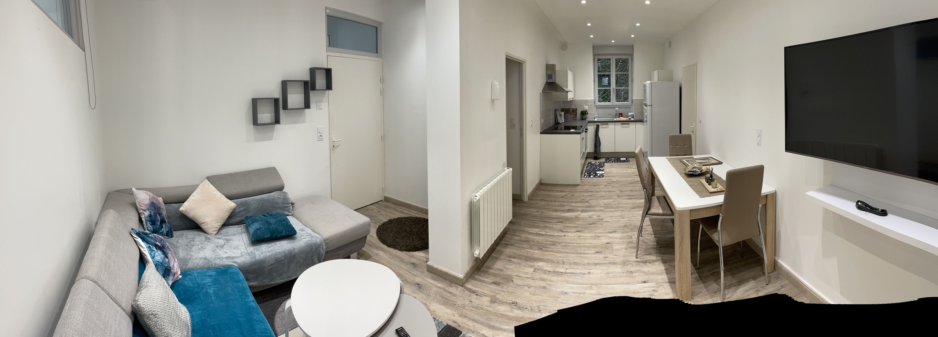 Een moderne woonkamer met een grijze bank, blauwe kussens, een ronde salontafel en een televisie aan de rechterkant.