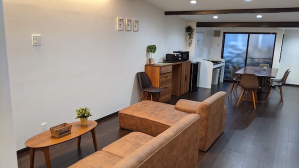 A modern living room with a sofa set, a dining table with chairs, a kitchen area, and a large window.