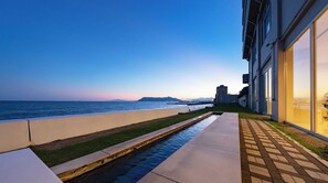 Un paseo marítimo con fuente de agua, vista al océano y un edificio con amplios ventanales.