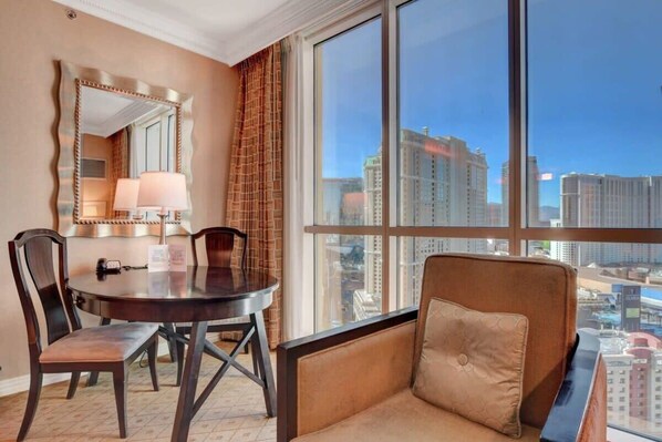 Ein Hotelzimmer mit einem runden Esstisch, zwei Stühlen, einer Couch mit Kissen, einem großen Spiegel und Blick auf die Stadt durch ein großes Fenster.
