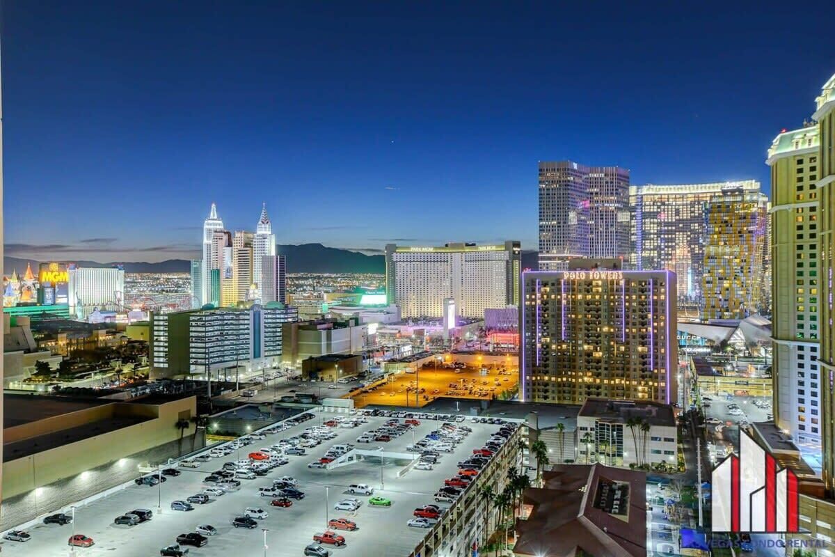 A cityscape at night with illuminated buildings, a parking lot, and a view of the mountains in the background.
