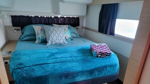 Una cama compacta con cubrecama y almohadas azules, una mesita de noche pequeña y una ventana con cortinas.