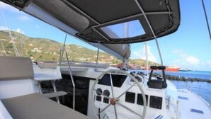 Interior de un barco con timón, paneles de control y vista al mar y tierra a lo lejos.