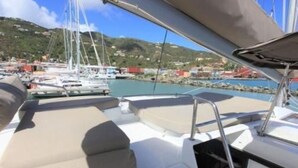Cubierta de barco con asientos y vista a un puerto deportivo con otras embarcaciones y un paisaje montañoso al fondo.