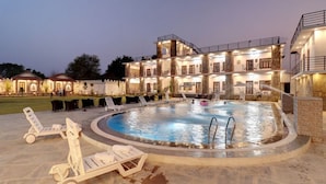 A large outdoor swimming pool surrounded by lounge chairs, with a multi-story building in the background. - Keshav Resort (Udaipur)