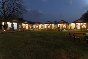 A row of small, stone-roofed buildings with arched doorways, arranged in a line across a grassy field. - Keshav Resort (Udaipur)