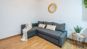 A modern living room with a grey sectional sofa, a white throw blanket, and patterned cushions. There's a round gold mirror on the wall and a small round table with a plant.