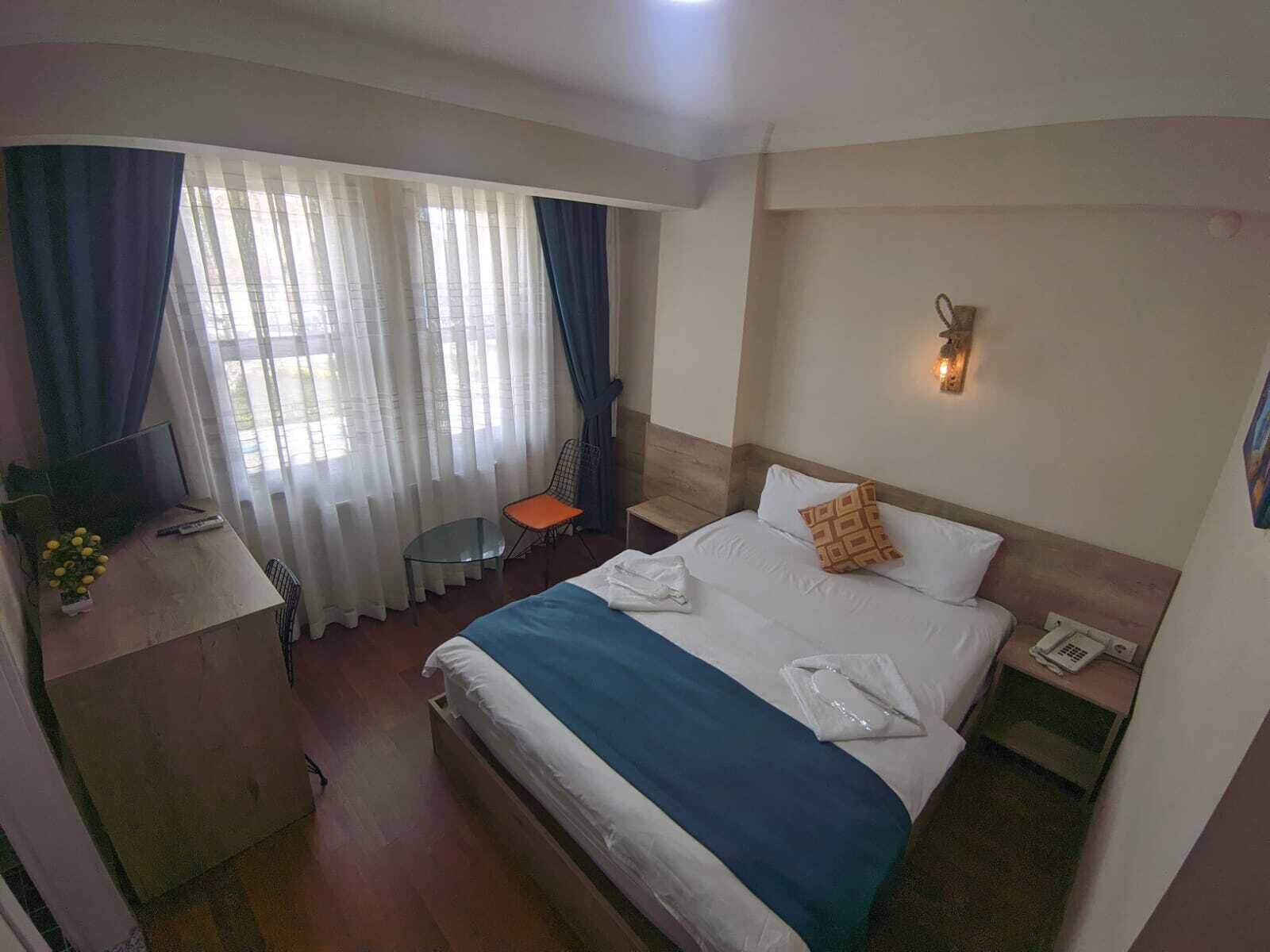 A hotel room with a bed, a desk with a chair, a small table, and a window with curtains.