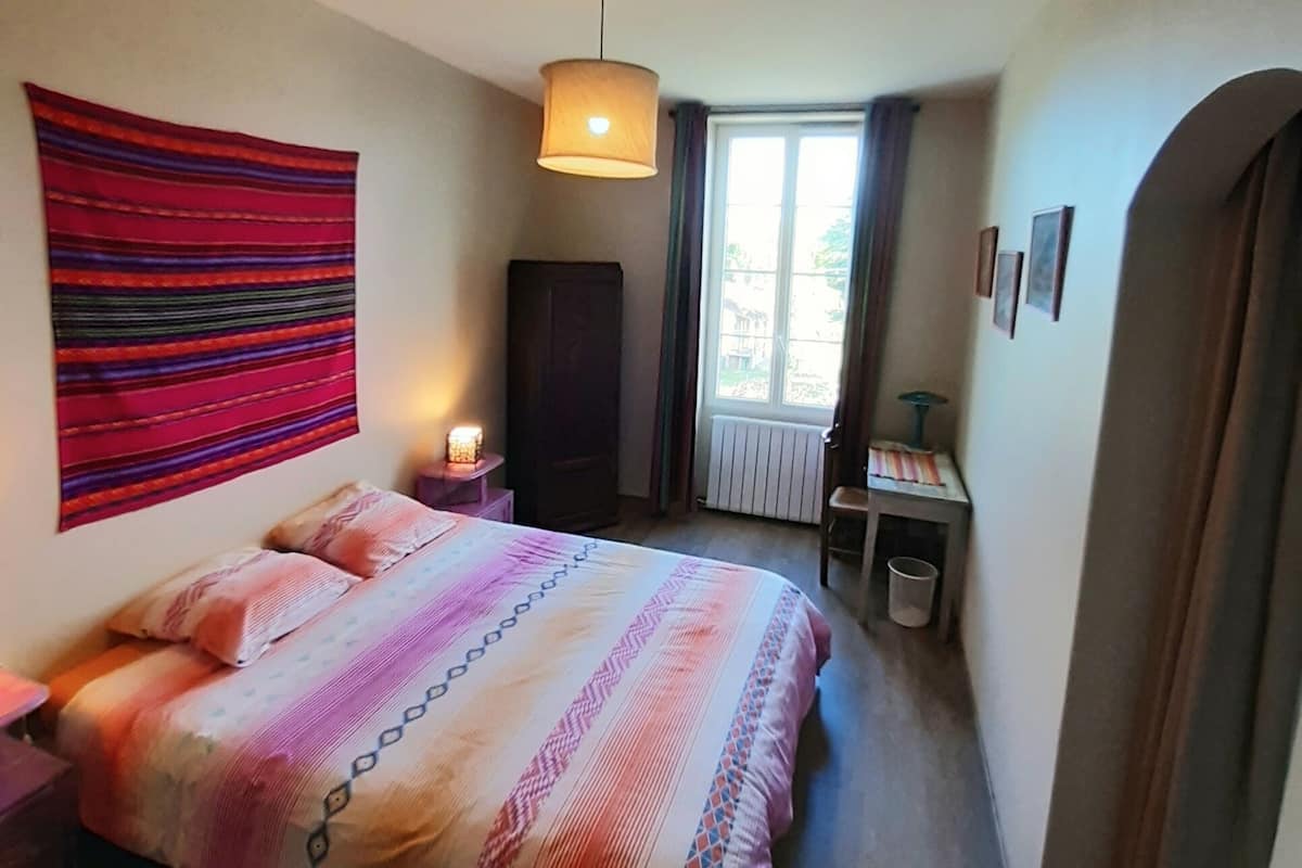 Une chambre à coucher avec un lit, une tapisserie murale, une fenêtre, un radiateur et un bureau avec une lampe.