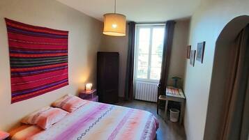 A bedroom with a bed, a hanging tapestry, a window, a radiator, and a desk with a lamp.