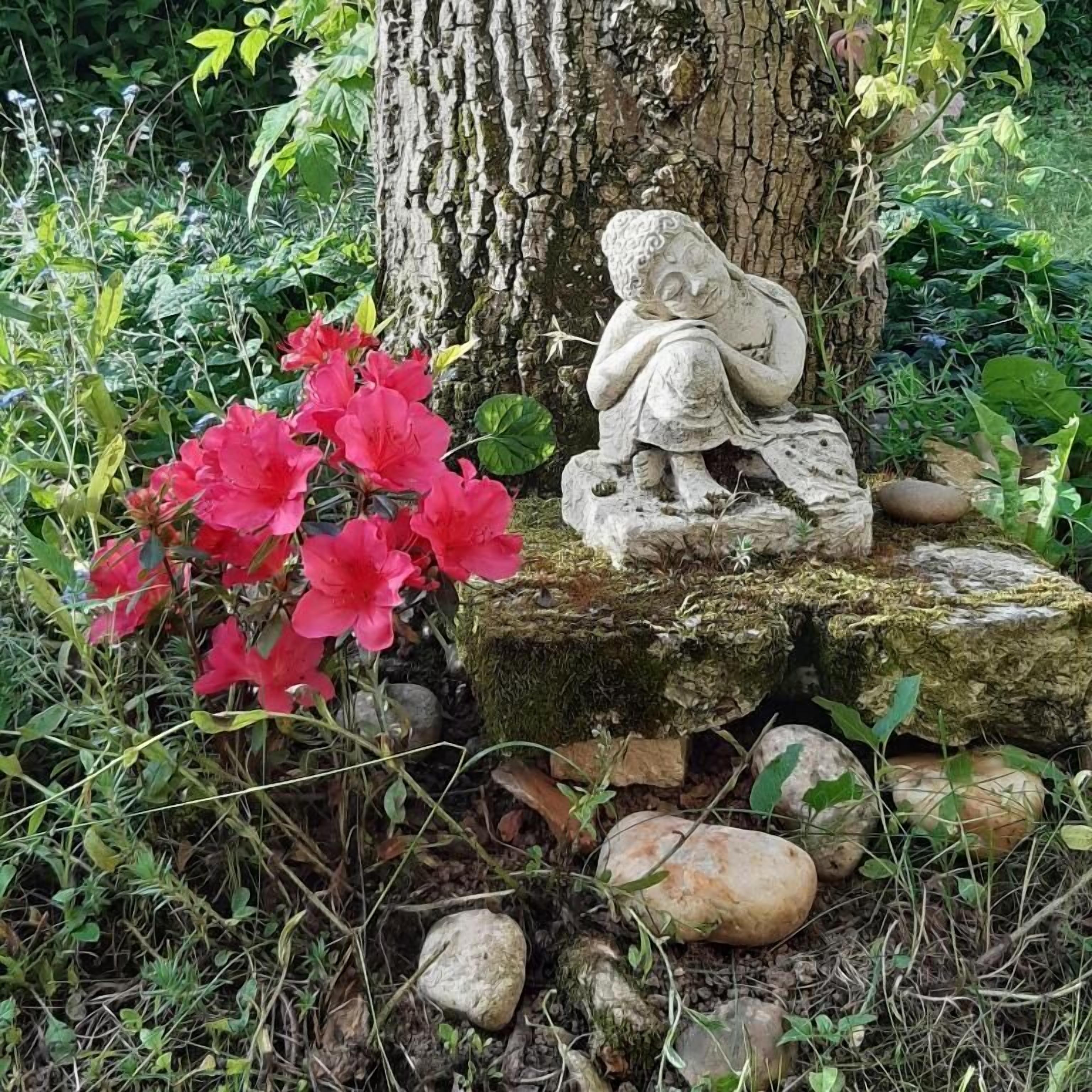 Un jardín con una estatua de piedra de una figura sentada sobre una roca, rodeado de follaje y flores rojas.