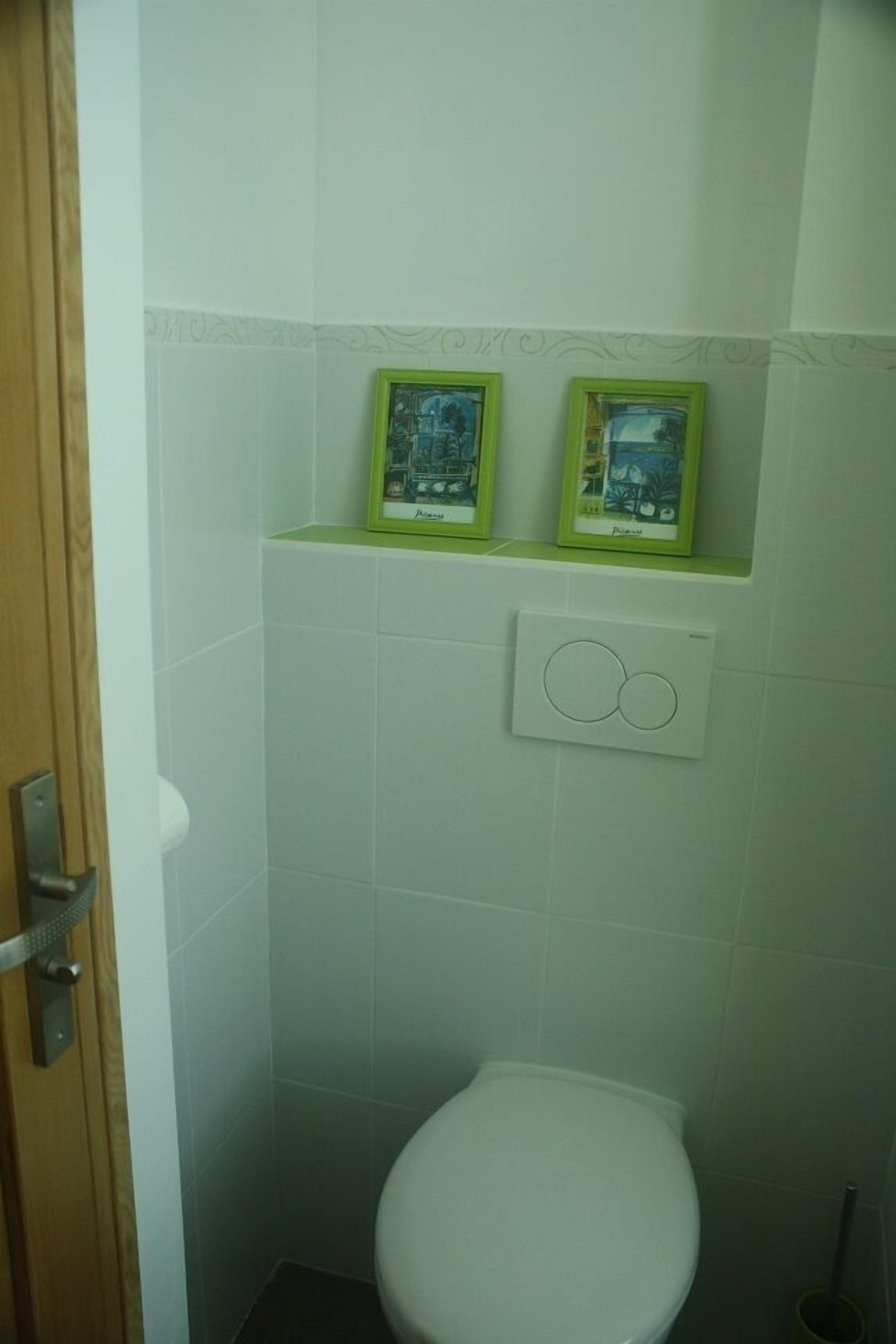 A small bathroom with a toilet, a wall-mounted light switch, and two framed pictures on the wall.