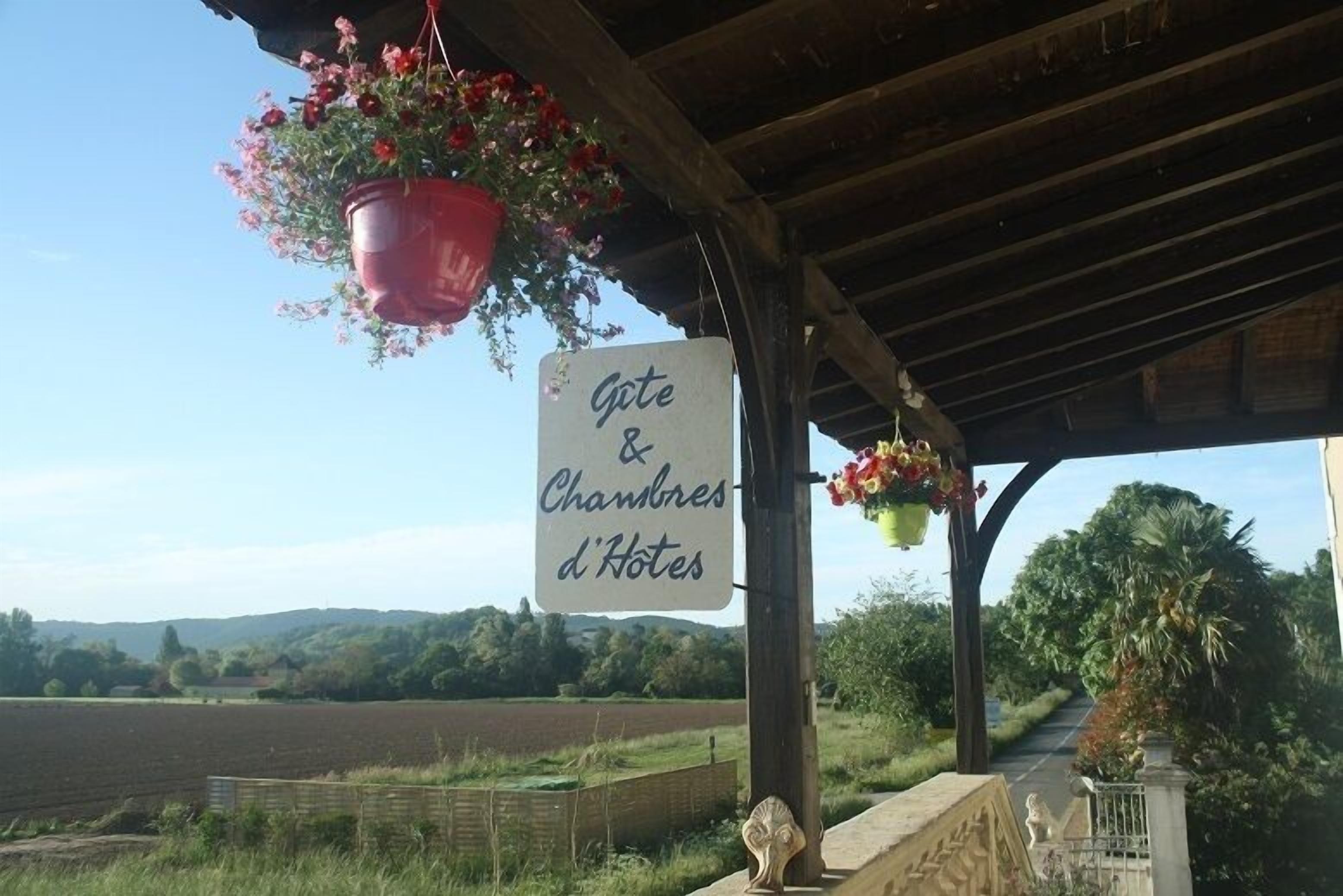 A sign for "Gîte & Chambres d'Hotes" with hanging flower baskets and a view of a rural landscape.