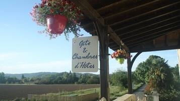 A sign for "Gîte & Chambres d'Hotes" with hanging flower baskets and a view of a rural landscape.