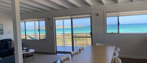 A spacious room with a large wooden table and white chairs, large windows with ocean views, and a balcony with a view of the beach.