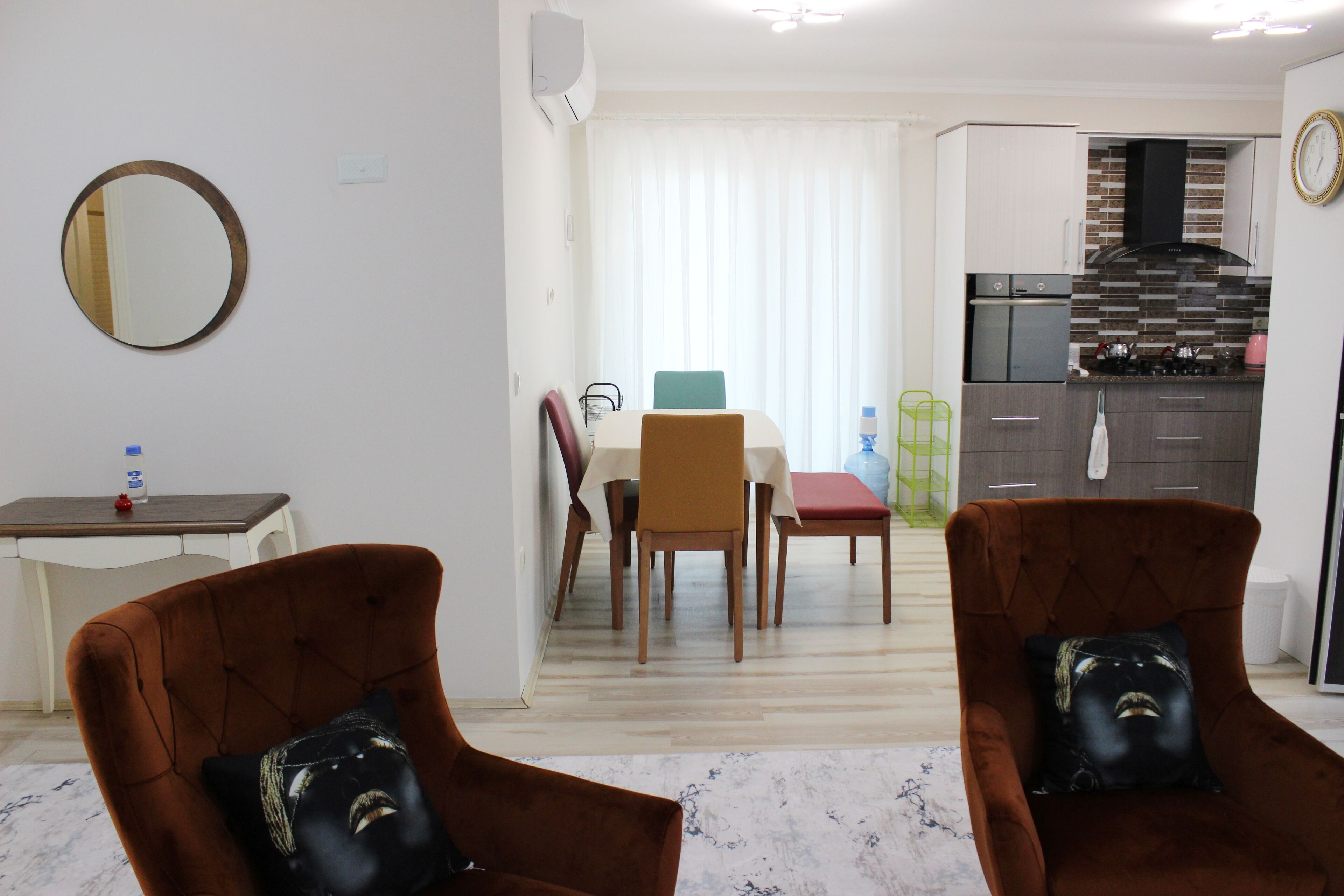 A modern living room with two brown armchairs, a dining area with a table and chairs, and a kitchen with a black range hood.
