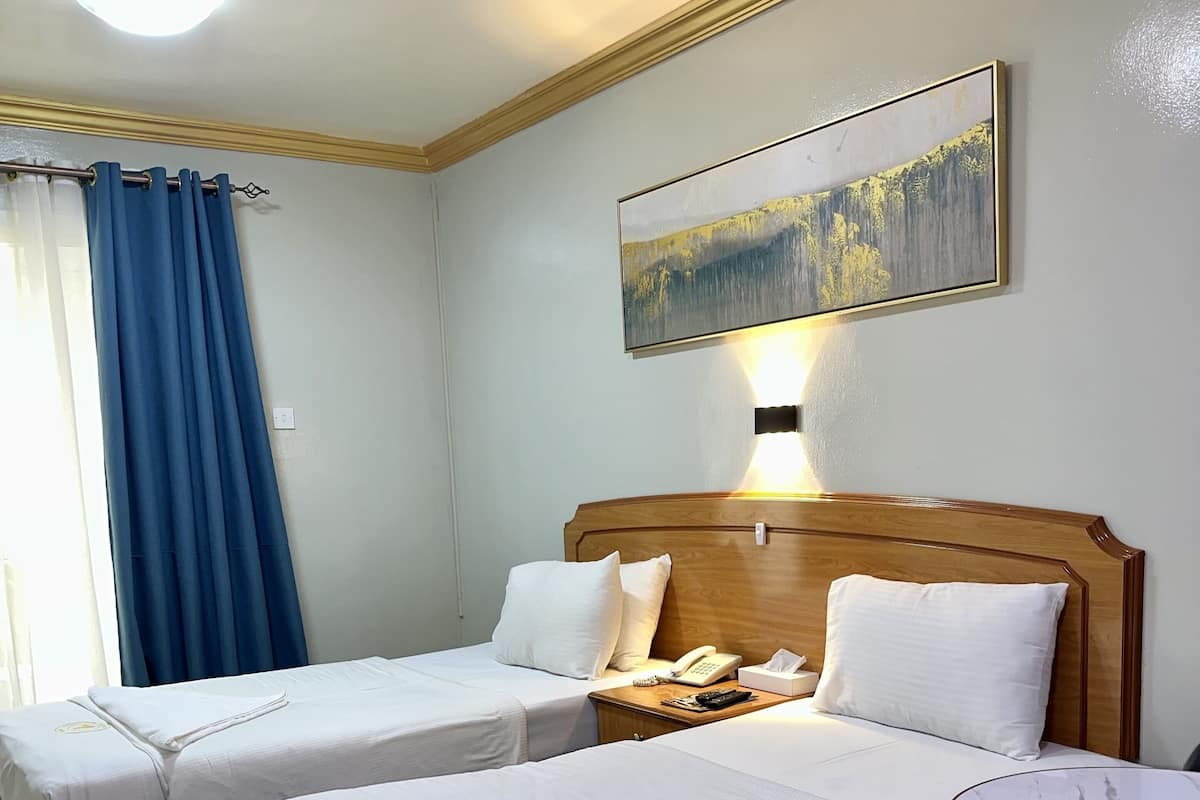A hotel room with two beds, a bedside table with a phone and tissues, a round table with a chair, and a painting on the wall.