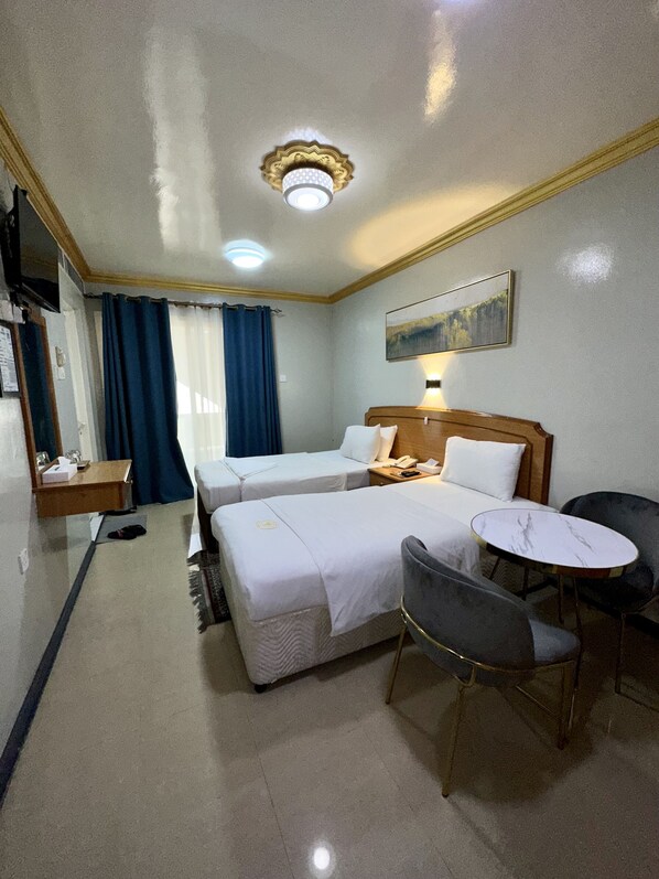 A hotel room with two single beds, a small round table, a chair, a desk with a telephone, and a painting on the wall.