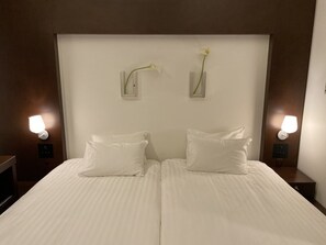Un lit bien fait, avec du linge de lit blanc, deux tables de chevet fixées au mur, chacune avec une lampe, et deux vases avec des fleurs blanches posés sur la tête de lit.