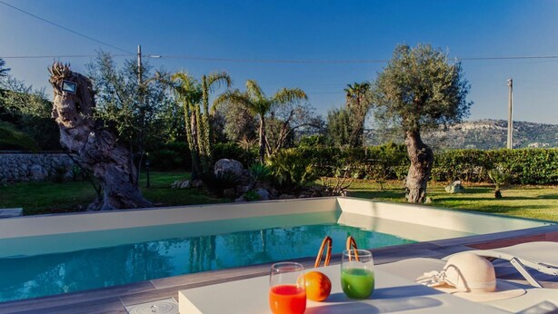 A swimming pool with a wooden deck, a lounge chair, and a table with drinks and a hat. - Villa Clara – Country Retreat (Siracusa)