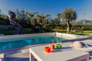 A swimming pool with a wooden deck, a lounge chair, and a table with drinks and a hat. - Villa Clara – A Getaway in the Countryside (Siracusa)