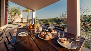 A wooden table set for breakfast with croissants, a pineapple, and a bottle of water, overlooking a scenic landscape. - Villa Clara – Country Retreat (Siracusa)