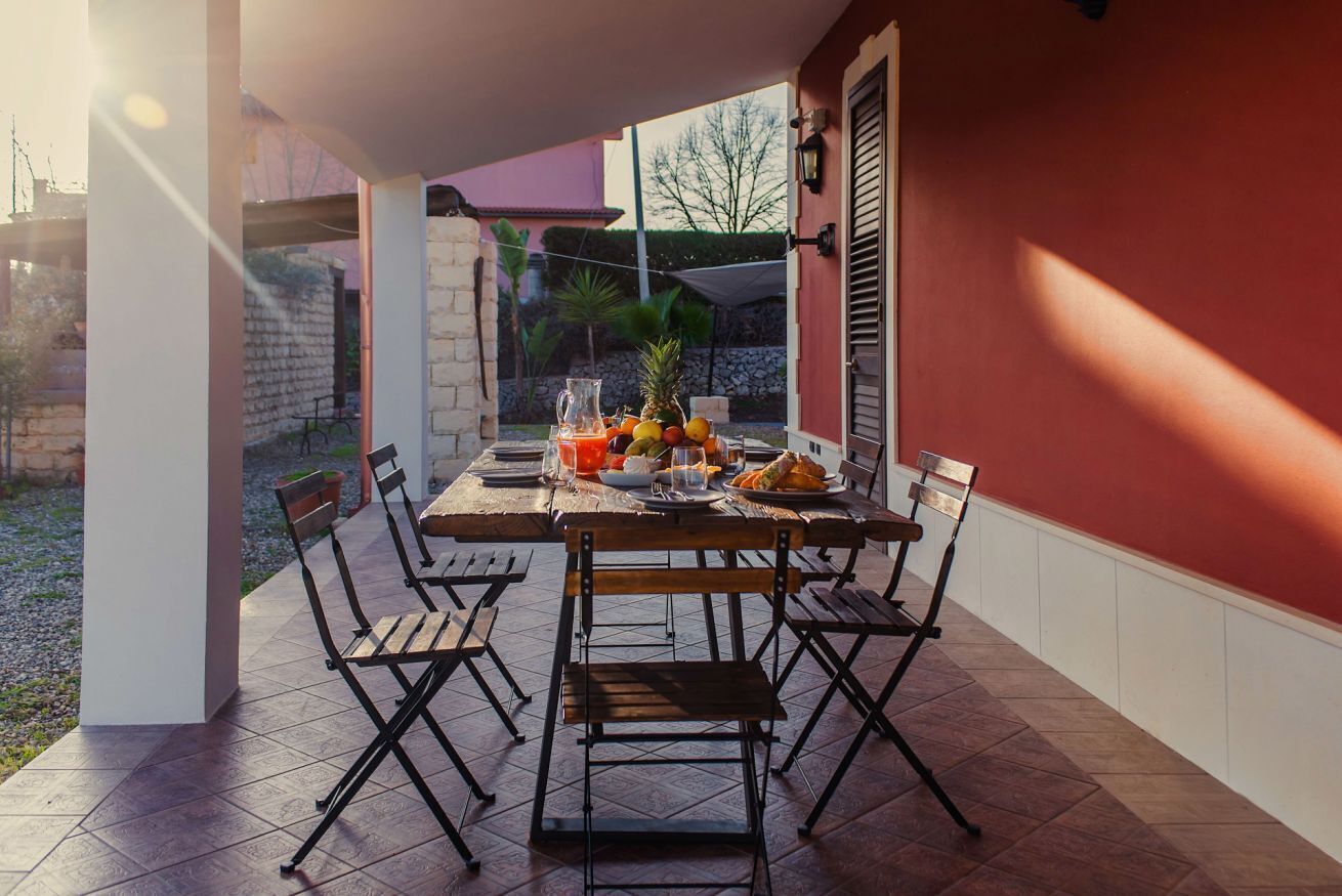 Eine überdachte Terrasse mit Tisch für vier Personen, umgeben von Stühlen und Blick auf Garten und Gebäude im Hintergrund.