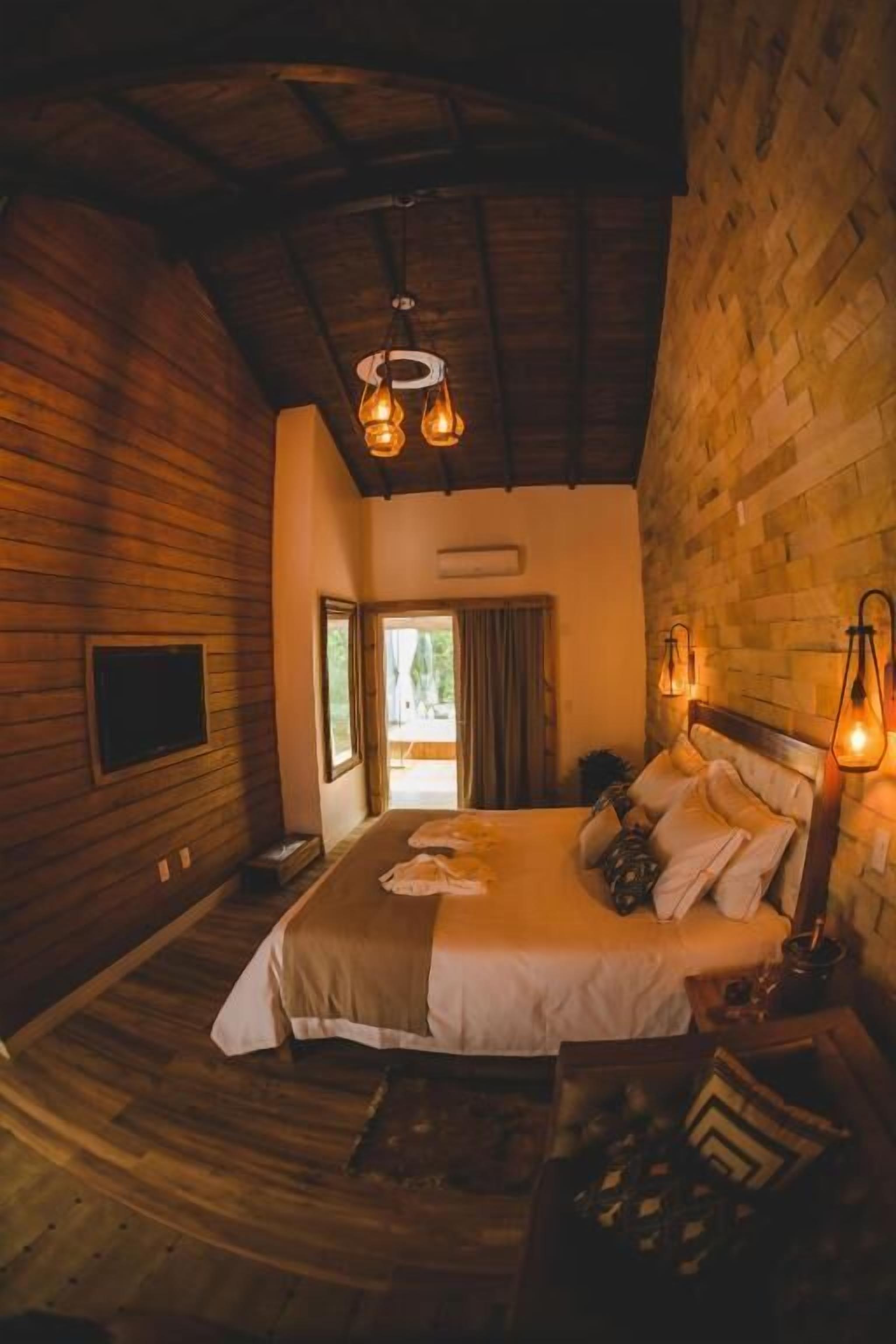 Quarto com teto de madeira, cama com roupa de cama branca e duas luminárias fixadas na parede.