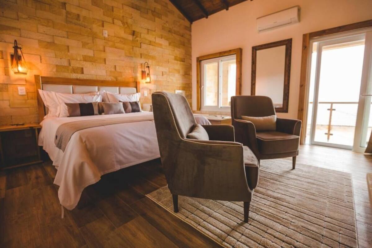 A bedroom with a brick wall, a bed with pillows, two chairs, a wooden floor, and a large window.