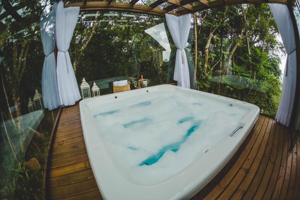 Uma banheira de hidromassagem em um deck de madeira com cortinas brancas, rodeada por vegetação exuberante.