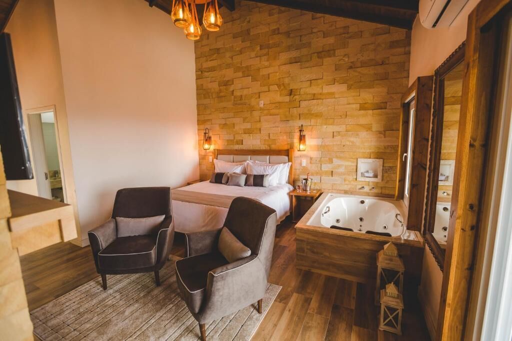 A bedroom with a wooden headboard, a large bed, two armchairs, a wooden nightstand, a white bathtub, and a mirror.